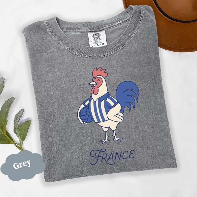 May include: A grey t-shirt with a cartoon rooster wearing a blue and white striped shirt and holding a ball. The word "France" is written in cursive below the rooster. The shirt is labeled "Comfort Colors".