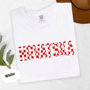 Może przedstawiać: Biała koszulka z napisem "HRVATSKA" w dużych, czerwono-białych kratkowanych literach z przodu. Koszulka wykonana jest z miękkiego materiału i ma okrągły dekolt.