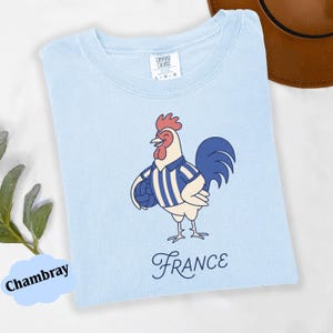 May include: Pale blue t-shirt featuring a cartoon rooster in a blue and white striped jersey, holding a football. The word "France" is written in cursive below the rooster. The shirt is labelled "Comfort Colors".