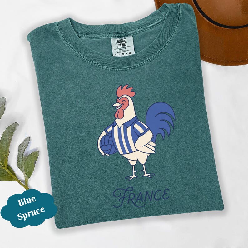 May include: Teal t-shirt with a cartoon rooster wearing a blue and white striped jersey and holding a football. The word "France" is written in a cursive font below the rooster. The shirt is a Comfort Colors brand.