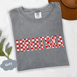 Może przedstawiać: Szara koszulka z napisem "HRVATSKA" w dużych, czerwono-białych kratkowanych literach. Koszulka ma okrągły dekolt i kr&oacute;tkie rękawy. Widoczna jest metka marki "COMFORT COLORS". Brązowy kapelusz jest częściowo widoczny w tle.