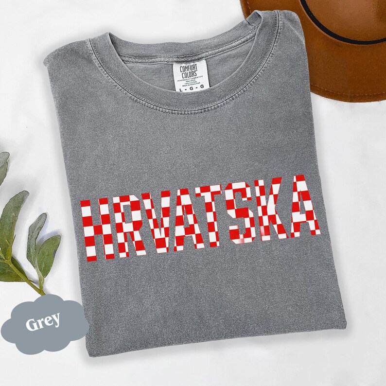 Może przedstawiać: Szara koszulka z napisem "HRVATSKA" w dużych, czerwono-białych kratkowanych literach. Koszulka ma okrągły dekolt i kr&oacute;tkie rękawy. Widoczna jest metka marki "COMFORT COLORS". Brązowy kapelusz jest częściowo widoczny w tle.