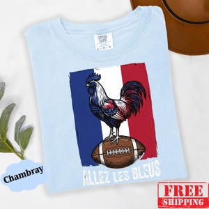 Puede incluir: Una camiseta azul claro con un gr&aacute;fico de un gallo de pie sobre un bal&oacute;n de f&uacute;tbol americano marr&oacute;n sobre el fondo de la bandera francesa. El texto "ALLEZ LES BLEUS" est&aacute; impreso en blanco debajo del bal&oacute;n. Tiene una etiqueta de Comfort Colors.
