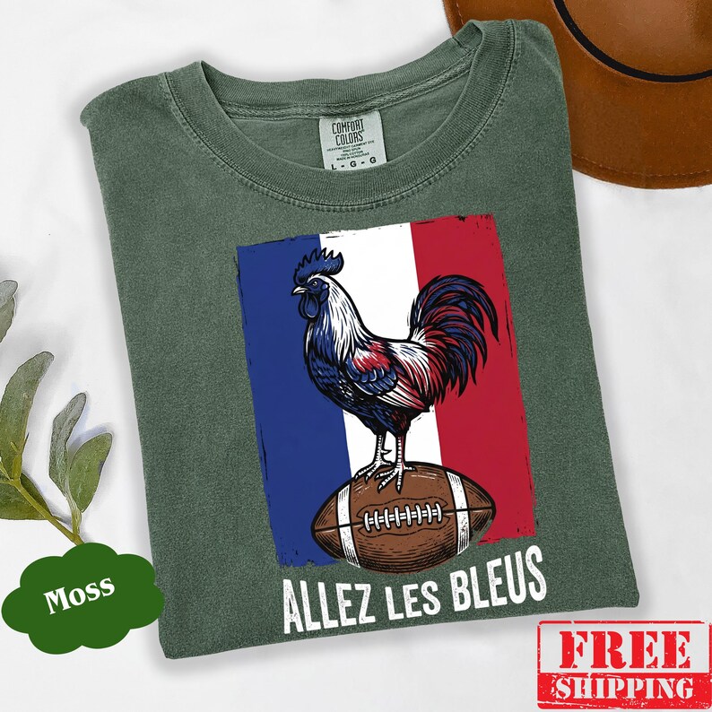 Puede incluir: Una camiseta de color verde musgo con un gr&aacute;fico de un gallo de colores azul, blanco y rojo de pie sobre un bal&oacute;n de f&uacute;tbol americano marr&oacute;n. Detr&aacute;s del gallo hay un dise&ntilde;o de bandera tricolor vertical. El texto "ALLEZ LES BLEUS" est&aacute; en blanco.