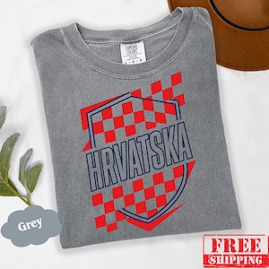 Pu&ograve; includere: Una t-shirt grigia a maniche corte con un motivo a scacchi rossi a forma di scudo e la scritta HRVATSKA in lettere blu scuro. La maglietta presenta un girocollo e un'etichetta del marchio Comfort Colors all'interno del colletto, taglia L.