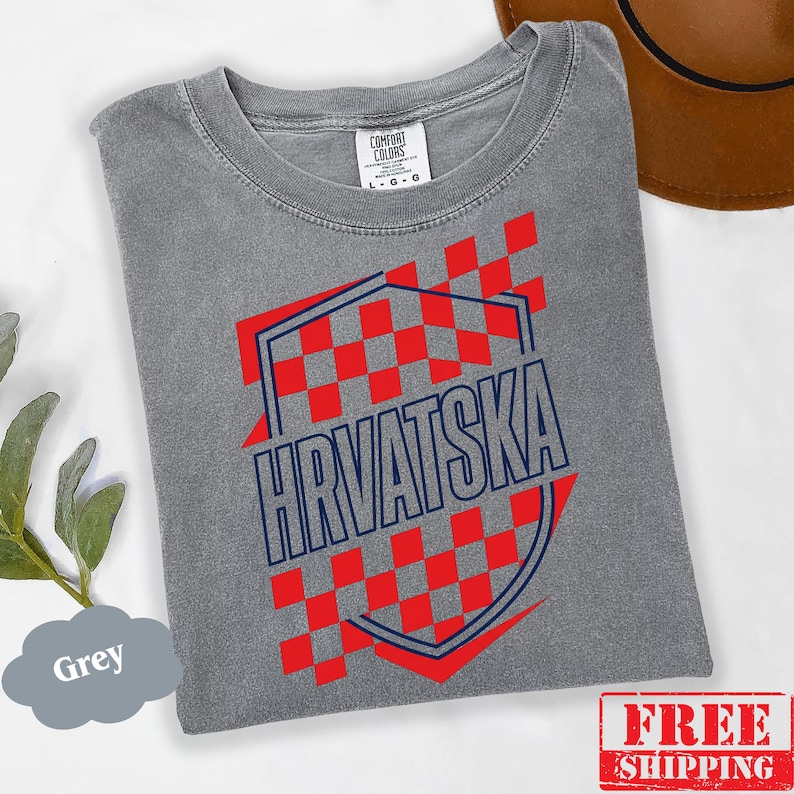Pu&ograve; includere: Una t-shirt grigia a maniche corte con un motivo a scacchi rossi a forma di scudo e la scritta HRVATSKA in lettere blu scuro. La maglietta presenta un girocollo e un'etichetta del marchio Comfort Colors all'interno del colletto, taglia L.