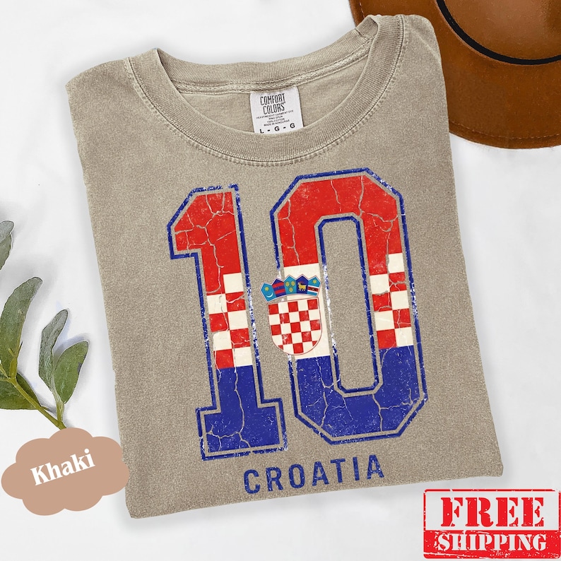 Puede incluir: Una camiseta de color caqui con un gr&aacute;fico grande y desgastado del n&uacute;mero 10 en rojo, blanco y azul, que incorpora el patr&oacute;n de la bandera croata y el escudo de armas. La palabra CROATIA est&aacute; impresa en azul debajo del gr&aacute;fico del n&uacute;mero 10.