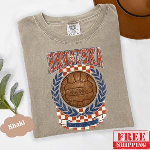 May include: A khaki-colored t-shirt featuring a graphic print with the text HRVATSKA in a distressed font. The design includes a brown vintage-style soccer ball centered within a blue laurel wreath, set against a red and white checkered background.
