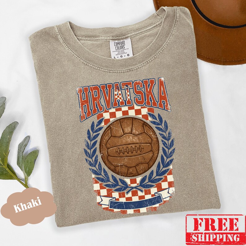 May include: A khaki-colored t-shirt featuring a graphic print with the text HRVATSKA in a distressed font. The design includes a brown vintage-style soccer ball centered within a blue laurel wreath, set against a red and white checkered background.