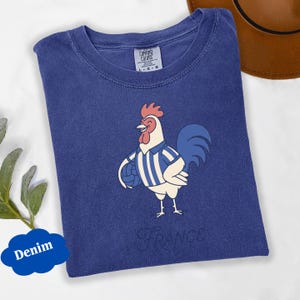 May include: A denim blue t-shirt with a cartoon rooster wearing a striped jersey and holding a ball. The word "France" is printed below the rooster. The shirt has a crew neck and the Comfort Colors brand label.