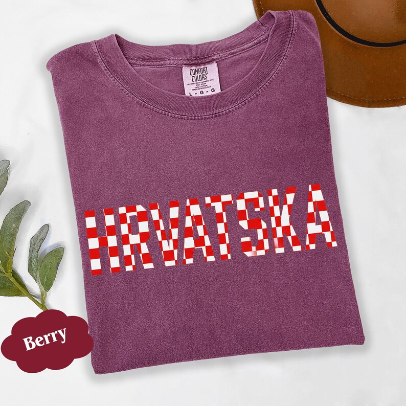Może przedstawiać: Koszulka w kolorze jagodowym z napisem "HRVATSKA" w czerwono-białą kratę. Koszulka jest marki Comfort Colors, a brązowy kapelusz jest widoczny w tle.