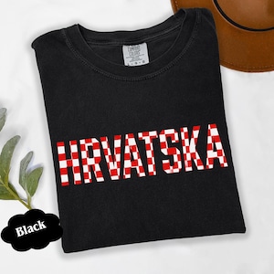 Może przedstawiać: Czarny t-shirt z napisem "HRVATSKA" w czerwono-białą kratę. Koszulka jest marki Comfort Colors i ma okrągły dekolt. W tle widoczny jest brązowy kapelusz.