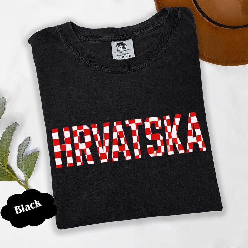 Może przedstawiać: Czarny t-shirt z napisem "HRVATSKA" w czerwono-białą kratę. Koszulka jest marki Comfort Colors i ma okrągły dekolt. W tle widoczny jest brązowy kapelusz.