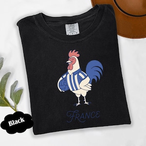 May include: Black t-shirt with a cartoon rooster wearing a blue and white striped jersey, holding a blue ball. The word "France" is written in cursive below the rooster. The shirt is labeled "Comfort Colors".