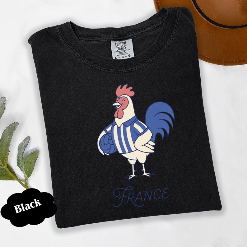 May include: Black t-shirt with a cartoon rooster wearing a blue and white striped jersey, holding a blue ball. The word "France" is written in cursive below the rooster. The shirt is labeled "Comfort Colors".