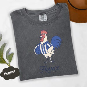 May include: A dark grey t-shirt with a cartoon rooster wearing a blue and white striped jersey, holding a ball. The word "France" is written in a cursive font below the rooster. The shirt has a Comfort Colors tag.