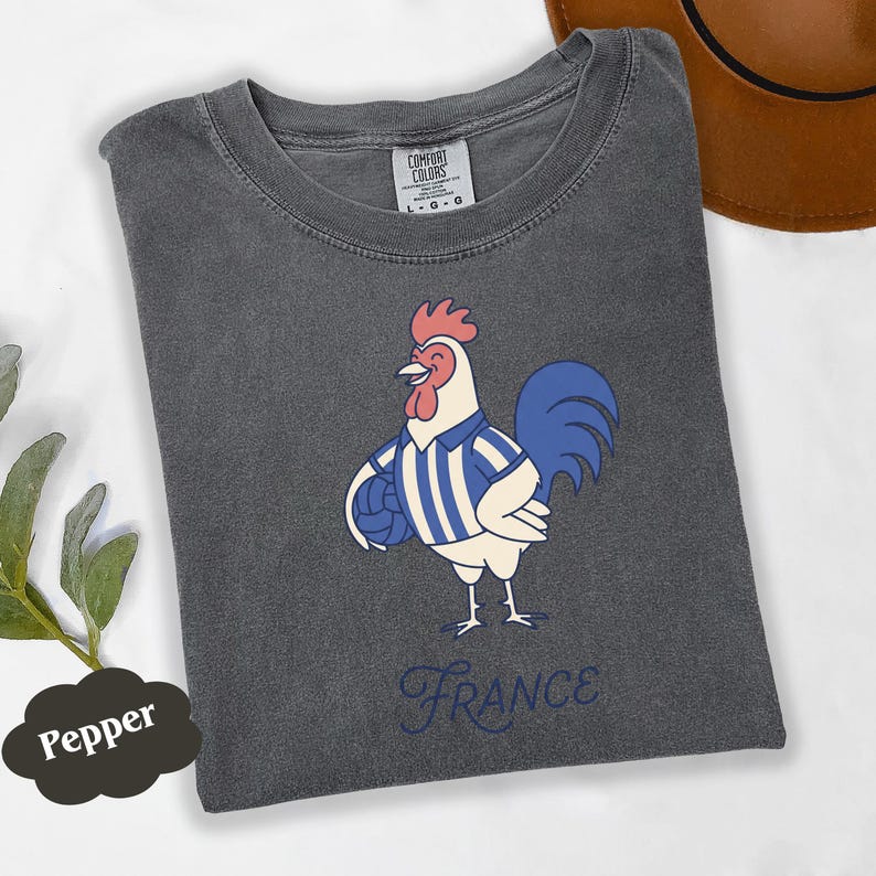 May include: A dark grey t-shirt with a cartoon rooster wearing a blue and white striped jersey, holding a ball. The word "France" is written in a cursive font below the rooster. The shirt has a Comfort Colors tag.