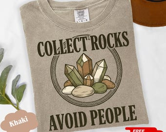 May include: A khaki crew neck t-shirt featuring a graphic of various crystals and stones inside a circular border. The text COLLECT ROCKS is printed above the graphic, and the text AVOID PEOPLE is printed below the graphic in dark brown ink.