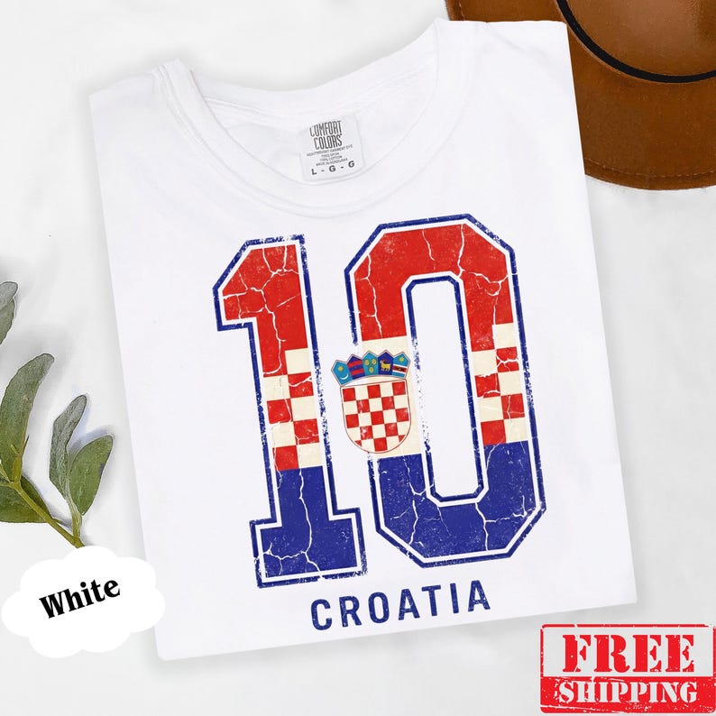 Puede incluir: Una camiseta blanca de manga corta con un gr&aacute;fico grande y desgastado del n&uacute;mero 10 en rojo y azul con un patr&oacute;n de cuadros. El escudo de armas croata est&aacute; centrado entre los d&iacute;gitos y la palabra CROATIA est&aacute; impresa debajo en azul.