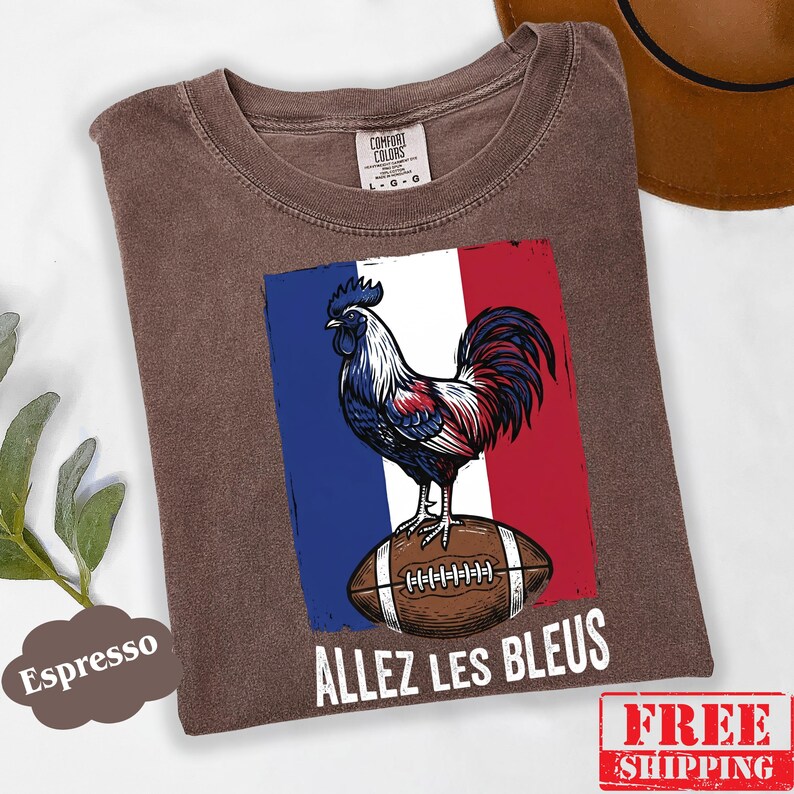 Puede incluir: Una camiseta marr&oacute;n de manga corta con un gr&aacute;fico de un gallo de pie sobre un bal&oacute;n de f&uacute;tbol americano marr&oacute;n sobre un fondo de bandera azul, blanca y roja. El texto "ALLEZ LES BLEUS" est&aacute; impreso en blanco debajo del gr&aacute;fico. Etiqueta Comfort Colors.