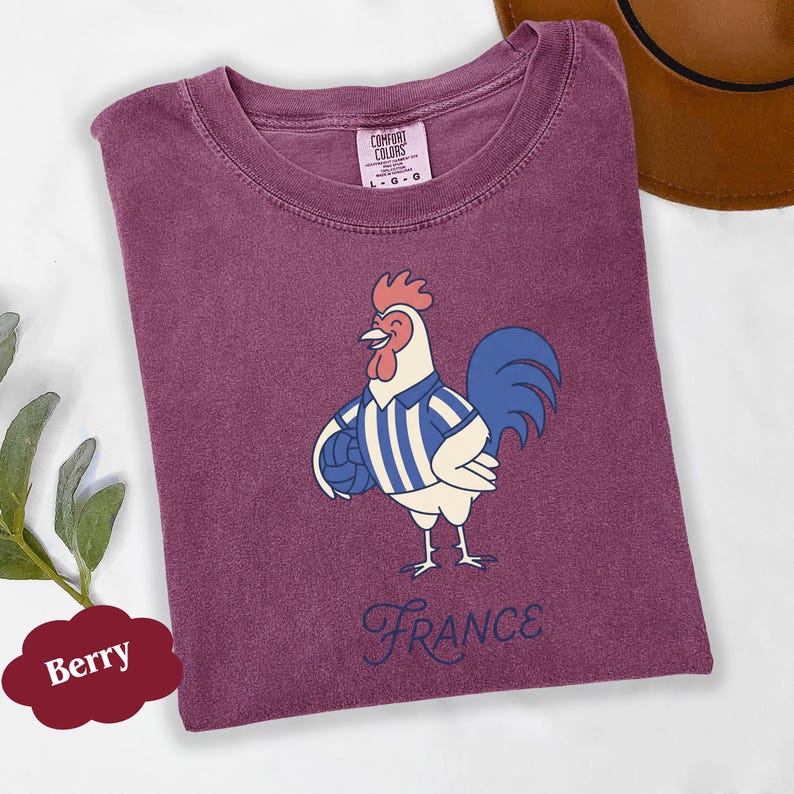 May include: Berry-coloured t-shirt with a cartoon rooster wearing a blue and white striped shirt and holding a football. The word "France" is written in a cursive font below the rooster. The shirt has a Comfort Colors tag.
