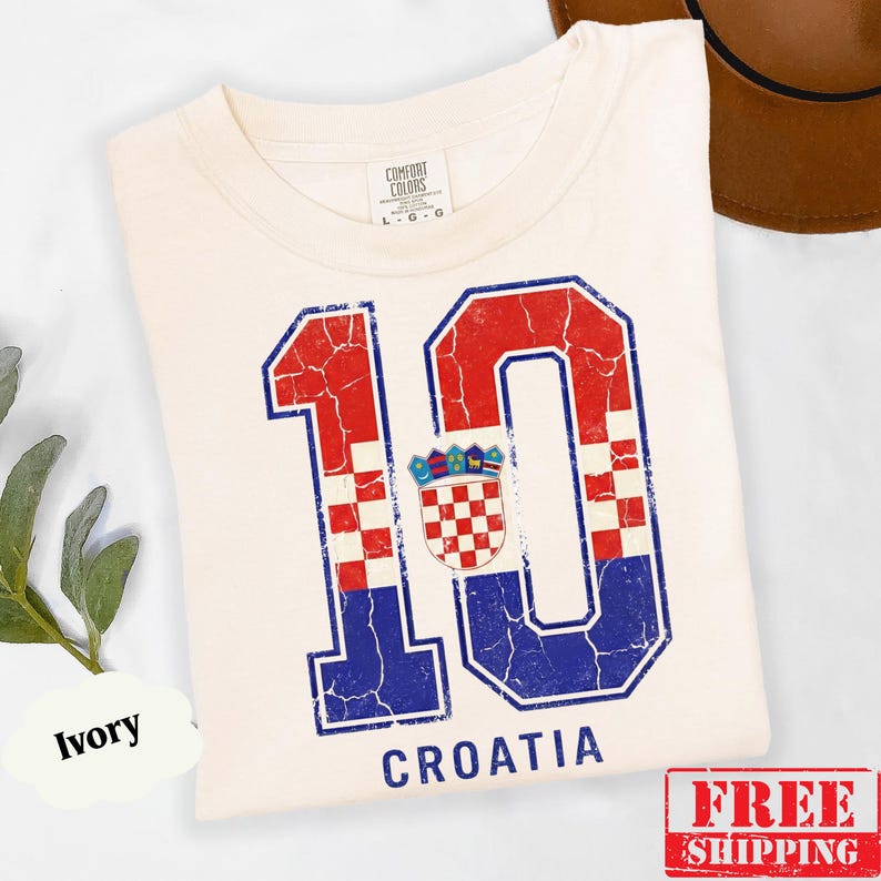 Puede incluir: Una camiseta de color marfil con un gr&aacute;fico grande y desgastado del n&uacute;mero 10, relleno con el patr&oacute;n de cuadros rojos y blancos de la bandera croata y detalles en azul. El escudo de armas de Croacia est&aacute; centrado entre los d&iacute;gitos, con la palabra CROATIA.