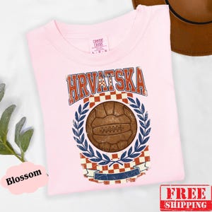 May include: A light pink t-shirt featuring a distressed graphic with the text HRVATSKA in blue and red. The design includes a brown vintage-style soccer ball centered above a red and white checkered pattern, framed by blue laurel leaves.