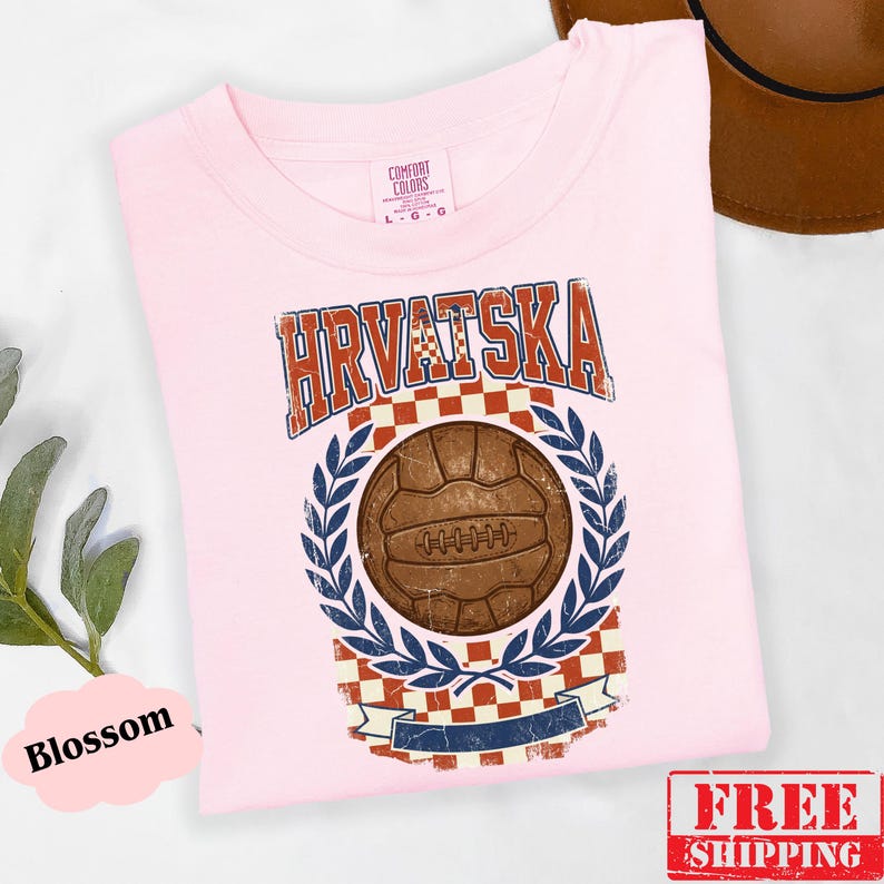 May include: A light pink t-shirt featuring a distressed graphic with the text HRVATSKA in blue and red. The design includes a brown vintage-style soccer ball centered above a red and white checkered pattern, framed by blue laurel leaves.