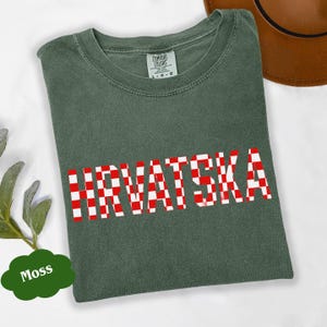 Może przedstawiać: Zielona koszulka w kolorze mchu z napisem "HRVATSKA" w czerwono-białą kratę. Koszulka ma okrągły dekolt i kr&oacute;tkie rękawy. Widoczna jest metka marki Comfort Colors.