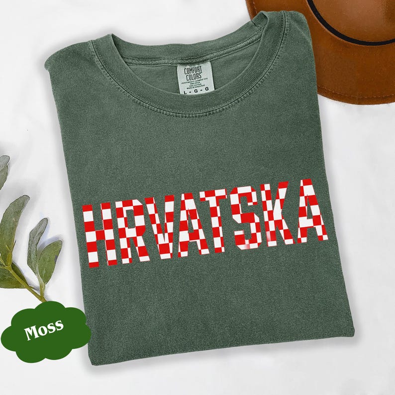 Może przedstawiać: Zielona koszulka w kolorze mchu z napisem "HRVATSKA" w czerwono-białą kratę. Koszulka ma okrągły dekolt i kr&oacute;tkie rękawy. Widoczna jest metka marki Comfort Colors.
