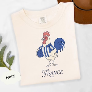May include: Ivory t-shirt with a cartoon rooster wearing a blue and white striped jersey, holding a ball. The word "France" is written in cursive below the rooster. The shirt has a Comfort Colors label.