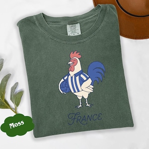 May include: A moss green t-shirt featuring a cartoon rooster in a blue and white striped jersey, holding a football. The word "France" is written in a cursive font below the rooster.