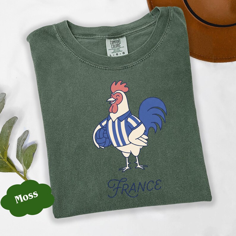 May include: A moss green t-shirt featuring a cartoon rooster in a blue and white striped jersey, holding a football. The word "France" is written in a cursive font below the rooster.