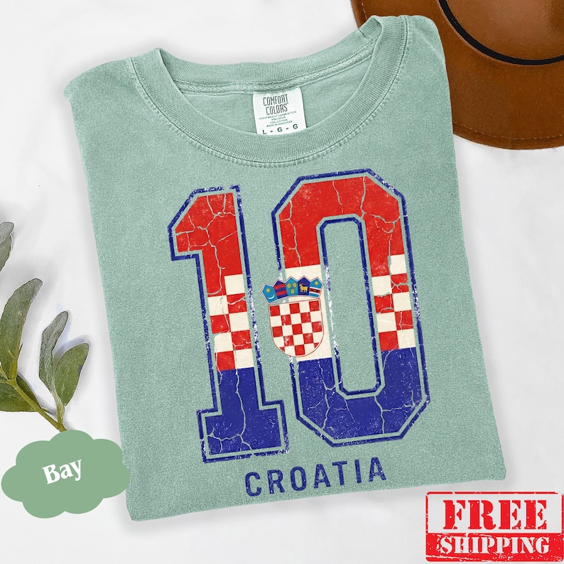 Puede incluir: Una camiseta de manga corta color verde salvia con un gr&aacute;fico grande y desgastado del n&uacute;mero 10 con un patr&oacute;n de cuadros rojos y blancos y detalles azules. El dise&ntilde;o incluye el escudo de armas croata y la palabra CROATIA impresa en azul.