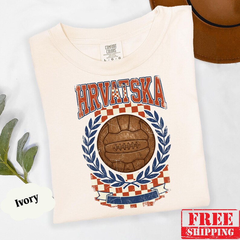 May include: An ivory t-shirt featuring a graphic print with the text HRVATSKA in red and blue. The design includes a brown vintage-style soccer ball centered within a blue laurel wreath, set against a red and white checkered background pattern.
