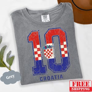 Puede incluir: Una camiseta gris de manga corta con un gr&aacute;fico grande y desgastado del n&uacute;mero 10 relleno con el patr&oacute;n de la bandera croata en rojo, blanco y azul, junto al escudo nacional. La palabra CROATIA est&aacute; impresa en azul debajo del n&uacute;mero 10.