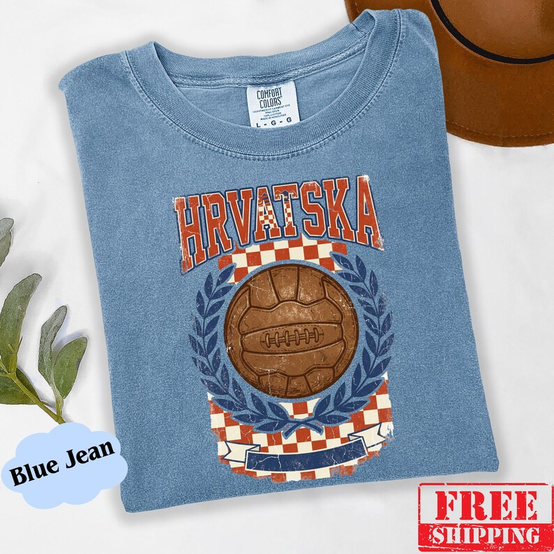 May include: A blue jean colored t-shirt featuring a vintage-style graphic with the text HRVATSKA in red letters. The design includes a brown soccer ball centered over a red and white checkered pattern, framed by blue laurel leaves and a banner.