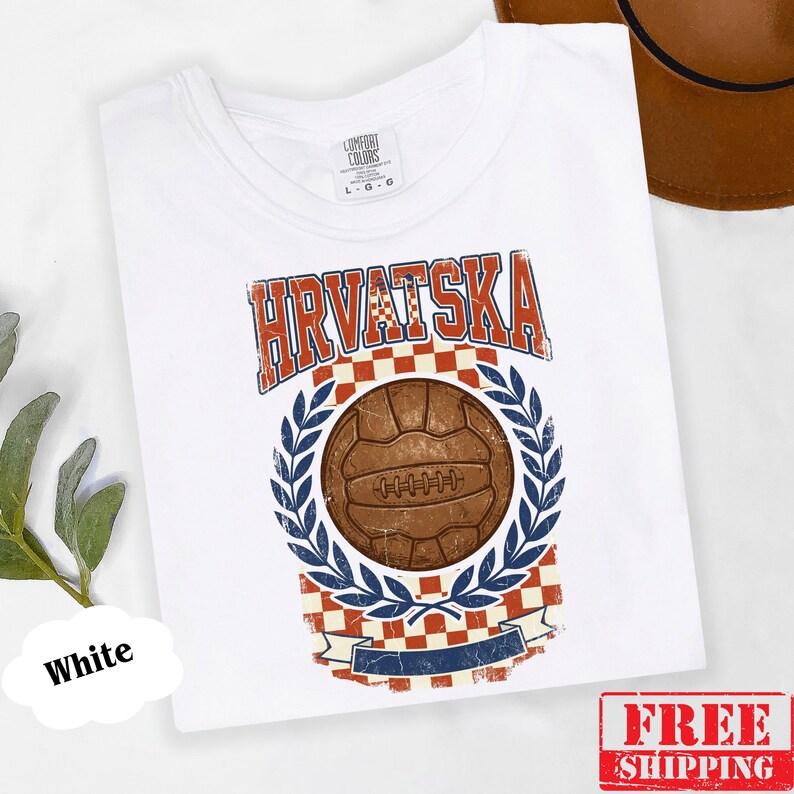 May include: A white t-shirt featuring a graphic print with the text HRVATSKA in red and blue letters. The design includes a brown vintage soccer ball centered above a red and white checkered pattern, framed by blue laurel leaves and a banner.