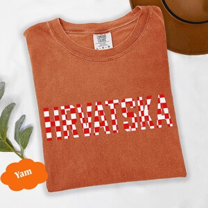 Może przedstawiać: Pomarańczowa koszulka z napisem "HRVATSKA" w czerwono-białą kratę. Koszulka jest złożona i ma metkę z napisem "Comfort Colors". W tle widoczny jest brązowy kapelusz.