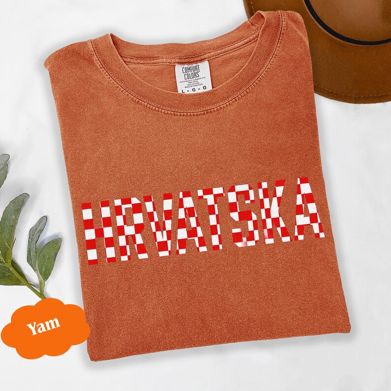 Może przedstawiać: Pomarańczowa koszulka z napisem "HRVATSKA" w czerwono-białą kratę. Koszulka jest złożona i ma metkę z napisem "Comfort Colors". W tle widoczny jest brązowy kapelusz.