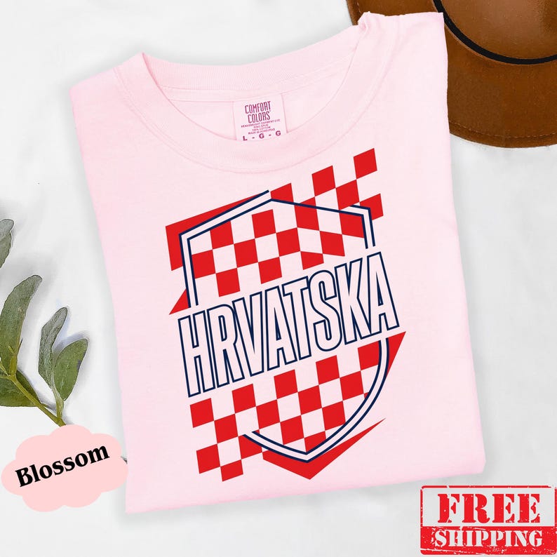 Pu&ograve; includere: Una t-shirt rosa chiaro con un design grafico a scacchi rossi e bianchi disposto a forma di scudo. La parola HRVATSKA &egrave; stampata in lettere maiuscole blu scuro al centro dello scudo. La maglietta presenta un classico girocollo.