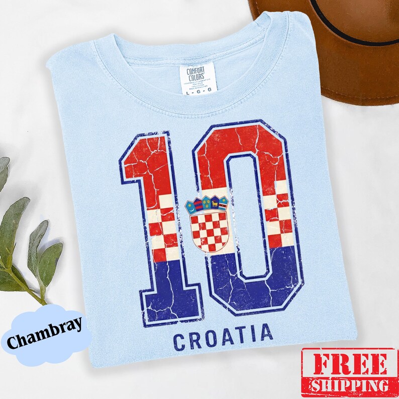 Puede incluir: Una camiseta azul claro con un gr&aacute;fico grande y desgastado del n&uacute;mero 10. El n&uacute;mero est&aacute; relleno con un patr&oacute;n de tablero de ajedrez rojo y blanco y una secci&oacute;n azul, incorporando el escudo de armas croata. La palabra CROATIA est&aacute; impresa debajo.