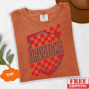 Pu&ograve; includere: Una t-shirt a maniche corte color ruggine con un motivo a scacchi rossi e bianchi e la parola HRVATSKA stampata in lettere maiuscole blu scuro all'interno di un contorno a forma di scudo. La maglietta ha un girocollo e un'etichetta Comfort Colors.