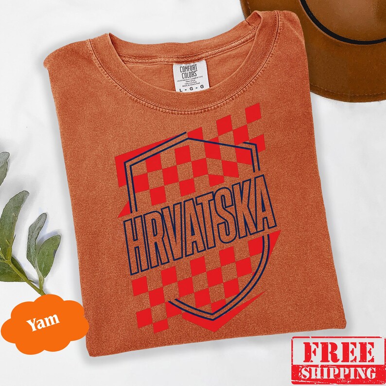 Pu&ograve; includere: Una t-shirt a maniche corte color ruggine con un motivo a scacchi rossi e bianchi e la parola HRVATSKA stampata in lettere maiuscole blu scuro all'interno di un contorno a forma di scudo. La maglietta ha un girocollo e un'etichetta Comfort Colors.