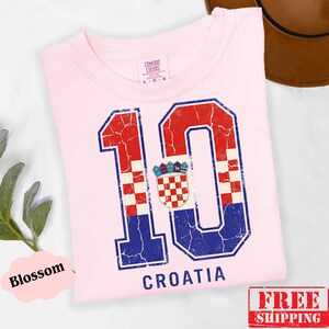 Puede incluir: Una camiseta rosa claro con un gr&aacute;fico grande y desgastado del n&uacute;mero 10, relleno con el patr&oacute;n de cuadros rojos y blancos de la bandera croata y detalles en azul. La palabra CROATIA est&aacute; impresa en azul debajo del gr&aacute;fico del n&uacute;mero 10.