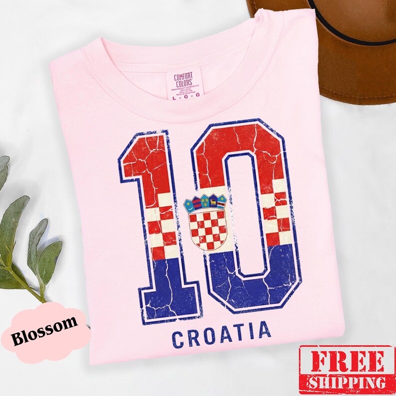 Puede incluir: Una camiseta rosa claro con un gr&aacute;fico grande y desgastado del n&uacute;mero 10, relleno con el patr&oacute;n de cuadros rojos y blancos de la bandera croata y detalles en azul. La palabra CROATIA est&aacute; impresa en azul debajo del gr&aacute;fico del n&uacute;mero 10.