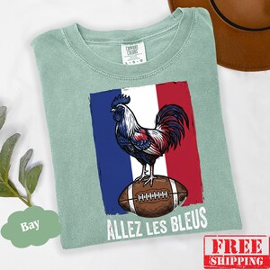 Puede incluir: Una camiseta verde salvia con un gr&aacute;fico de un gallo de pie sobre un bal&oacute;n de f&uacute;tbol americano marr&oacute;n sobre un fondo de bandera azul, blanca y roja. El texto "ALLEZ LES BLEUS" est&aacute; impreso en blanco debajo del bal&oacute;n. Etiqueta Comfort Colors.