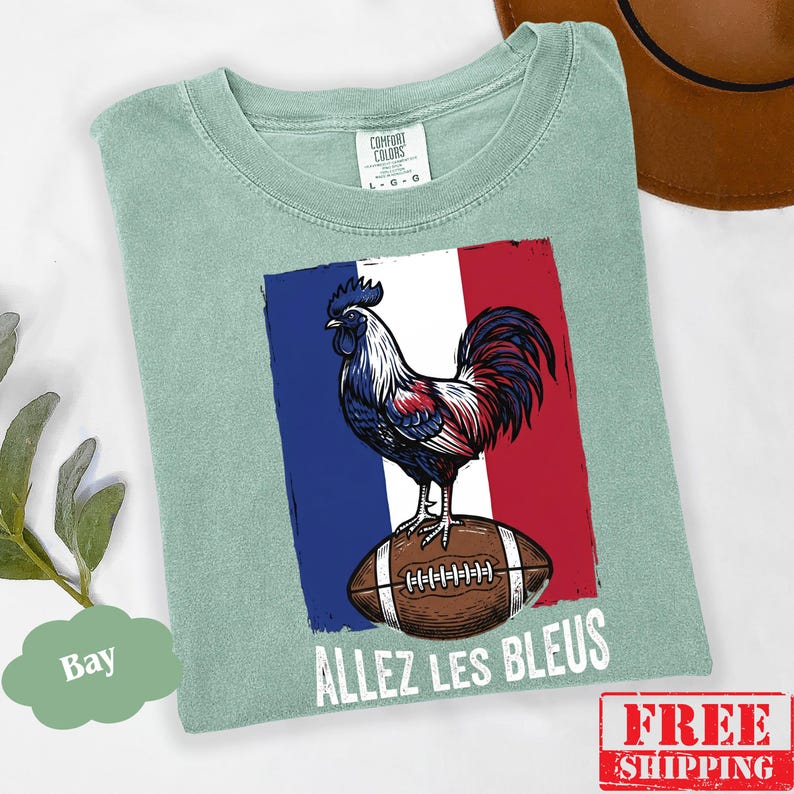 Puede incluir: Una camiseta verde salvia con un gr&aacute;fico de un gallo de pie sobre un bal&oacute;n de f&uacute;tbol americano marr&oacute;n sobre un fondo de bandera azul, blanca y roja. El texto "ALLEZ LES BLEUS" est&aacute; impreso en blanco debajo del bal&oacute;n. Etiqueta Comfort Colors.