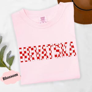 Może przedstawiać: Jasnor&oacute;żowa koszulka z napisem "HRVATSKA" dużymi, czerwono-białymi szachownicami. Koszulka jest złożona i ma metkę z napisem "Comfort Colors".
