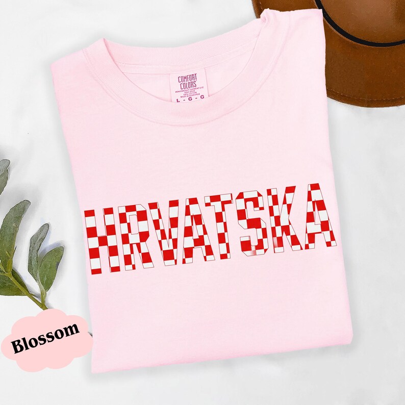 Może przedstawiać: Jasnor&oacute;żowa koszulka z napisem "HRVATSKA" dużymi, czerwono-białymi szachownicami. Koszulka jest złożona i ma metkę z napisem "Comfort Colors".